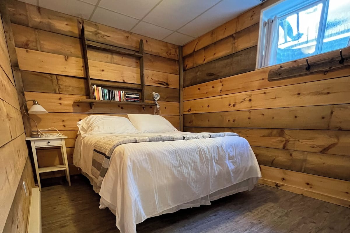A wooden cabin room with a bed, bedside table, lamp, and bookshelves.