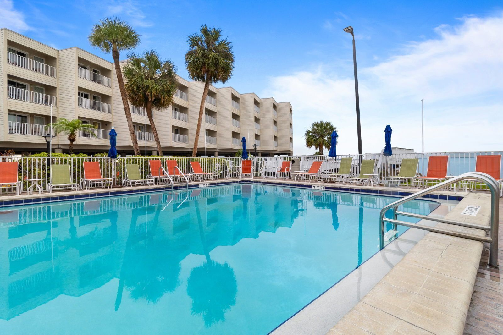 A swimming pool with lounge chairs and umbrellas, surrounded by a fence, with a hotel building in the background.