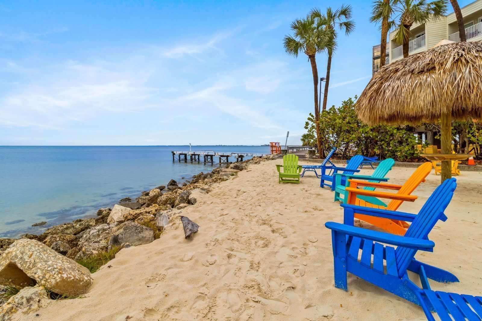 A beach with colorful Adirondack chairs, a thatched-roof structure, and a pier extending into the water.