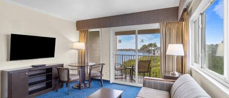 A hotel room with a flat-screen TV, a dining table with chairs, a sofa, a small table, and a balcony with a view of the water.