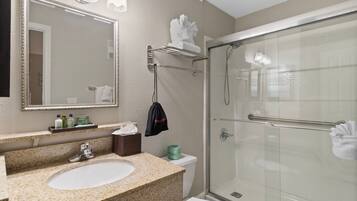 A bathroom with a granite countertop sink, a mirror with lights, a glass shower enclosure, and a towel rack.