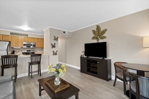 A modern living room with a flat-screen TV, dark wood furniture, a kitchen with wooden cabinets, and decorative wall art.