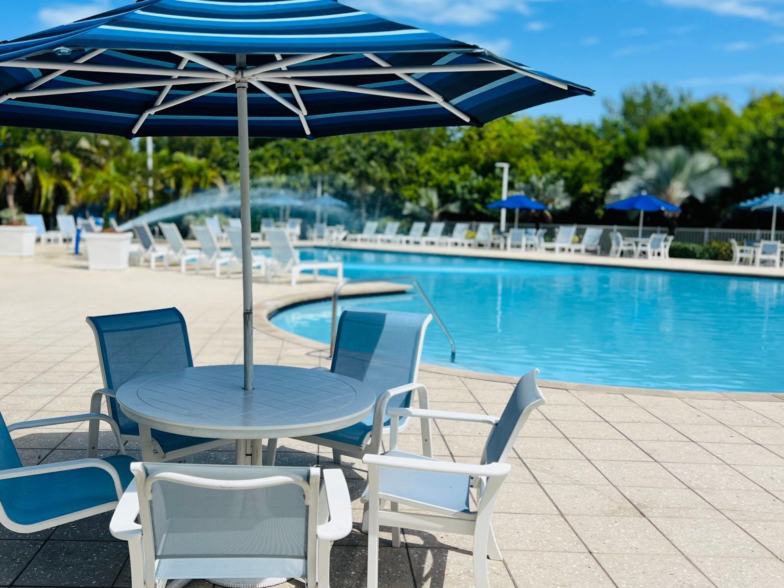 A poolside area with a round table and chairs, a blue umbrella, and lounge chairs.