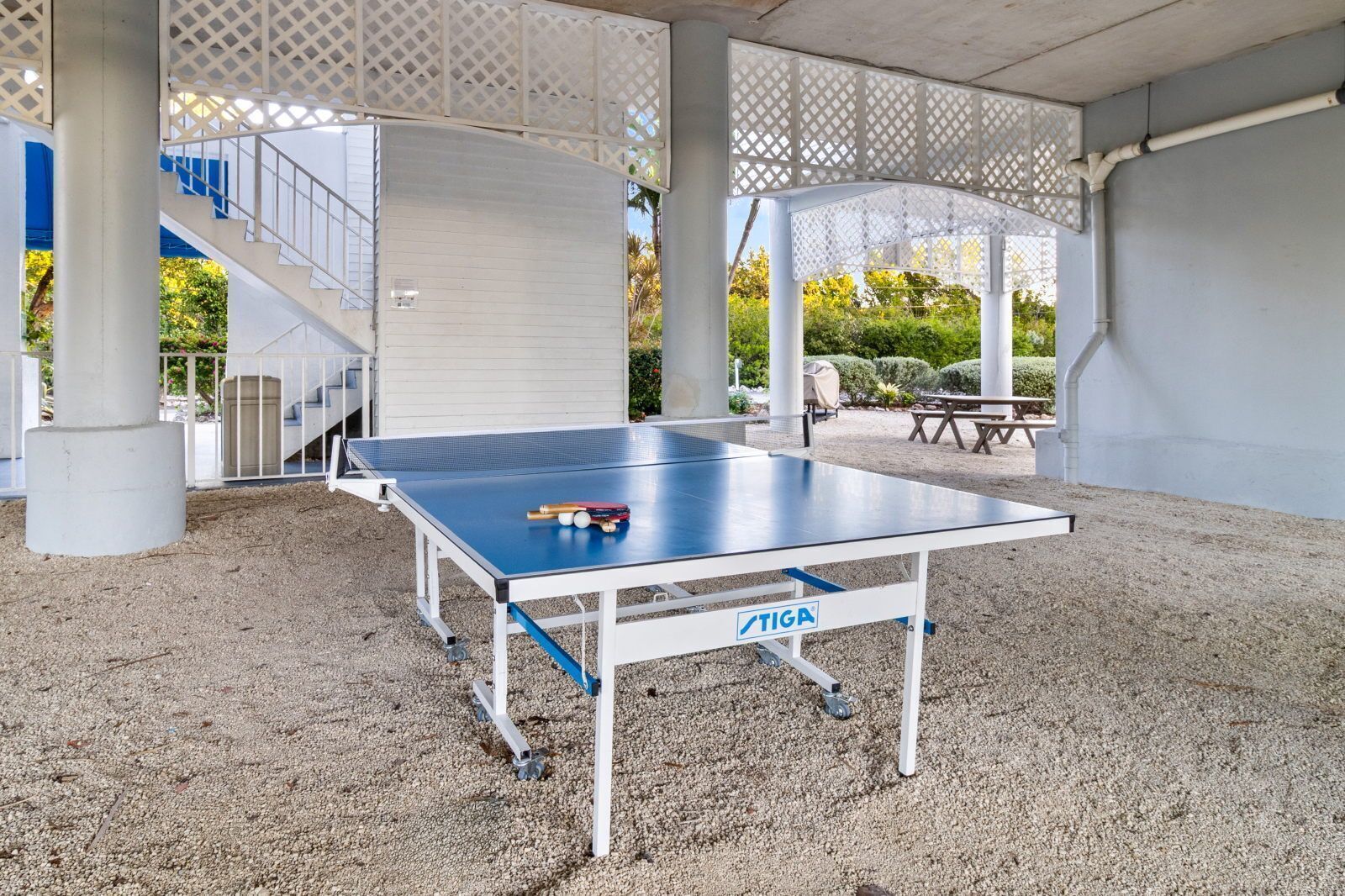 An outdoor ping pong table with a TIGA logo, surrounded by gravel, under a covered area with white lattice panels and a staircase.