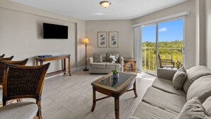 A spacious living room with a sofa, chairs, a coffee table, a flat-screen TV, and a balcony with a view.