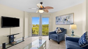 A living room with a flat-screen TV, a ceiling fan, a glass coffee table, a sofa, a chair, a lamp, and a balcony with a view.