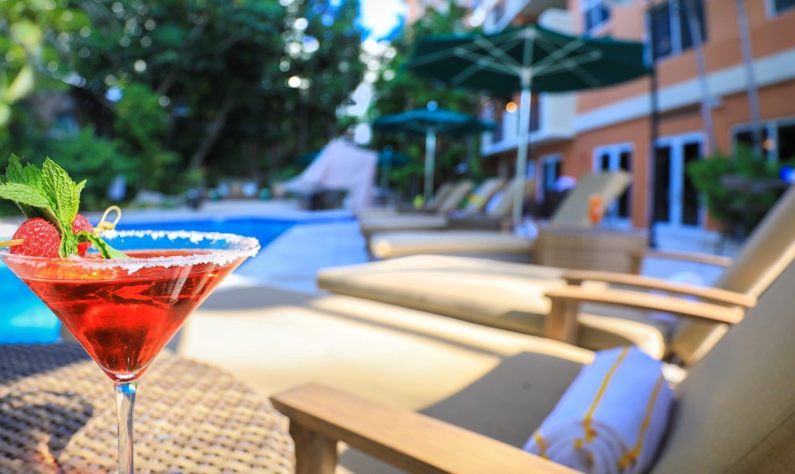 Une scène au bord d’une piscine, avec un cocktail servi dans un verre à martini, des chaises longues et un bâtiment en arrière-plan.