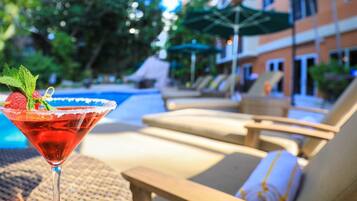 Une scène au bord d’une piscine, avec un cocktail servi dans un verre à martini, des chaises longues et un bâtiment en arrière-plan.