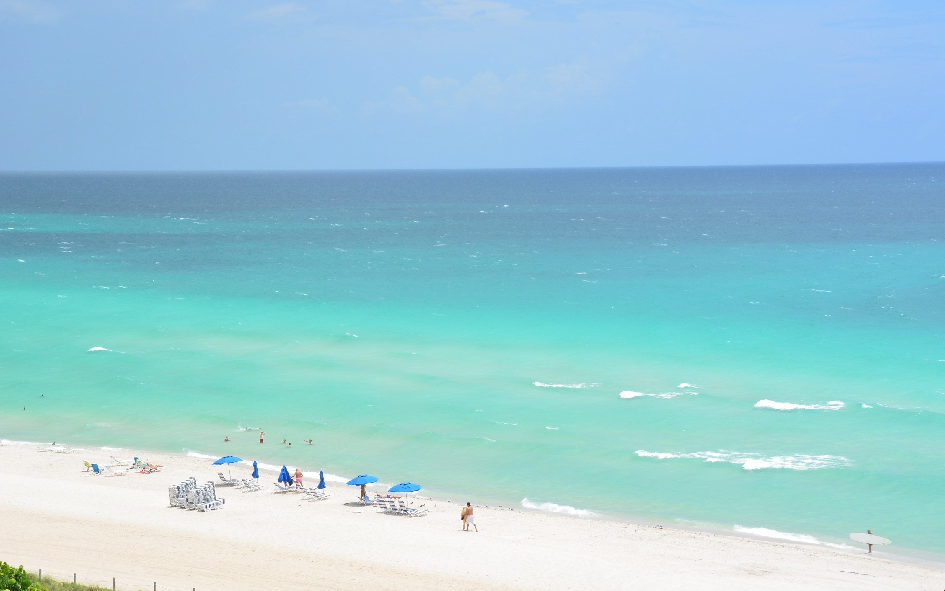 A beach with clear turquoise water, a sandy shore, and several blue umbrellas.