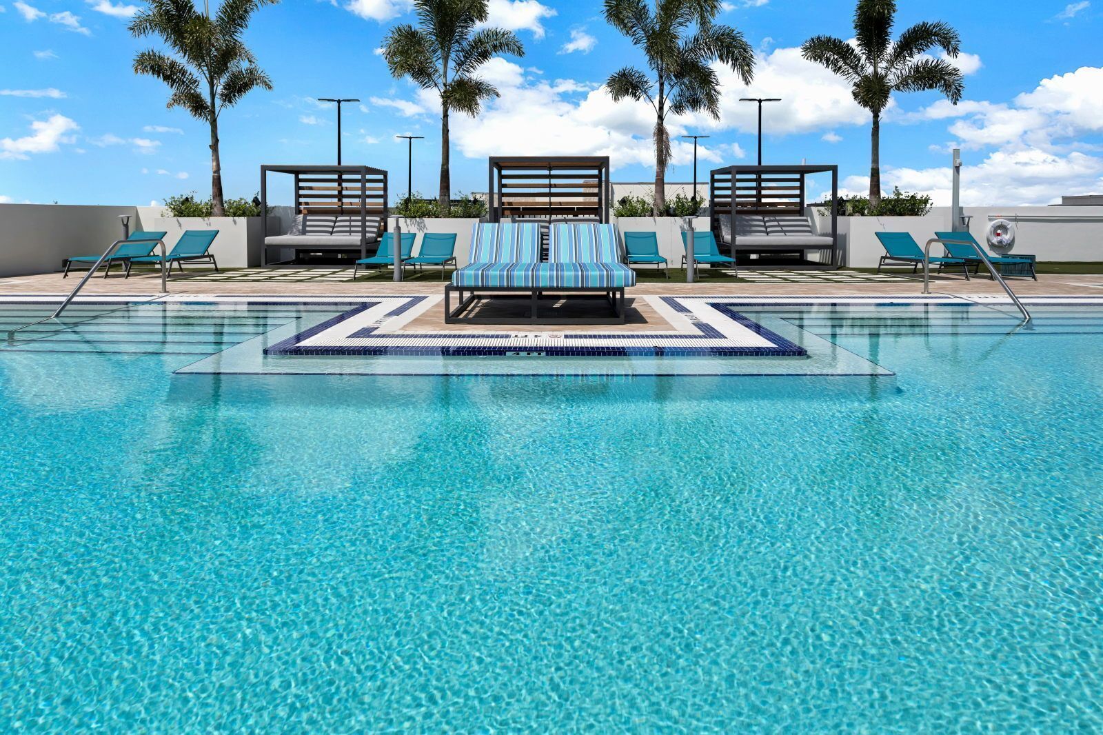 A clear blue pool with lounge chairs and a sunbed, surrounded by palm trees and a clear sky.