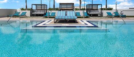 A clear blue pool with lounge chairs and a sunbed, surrounded by palm trees and a clear sky.