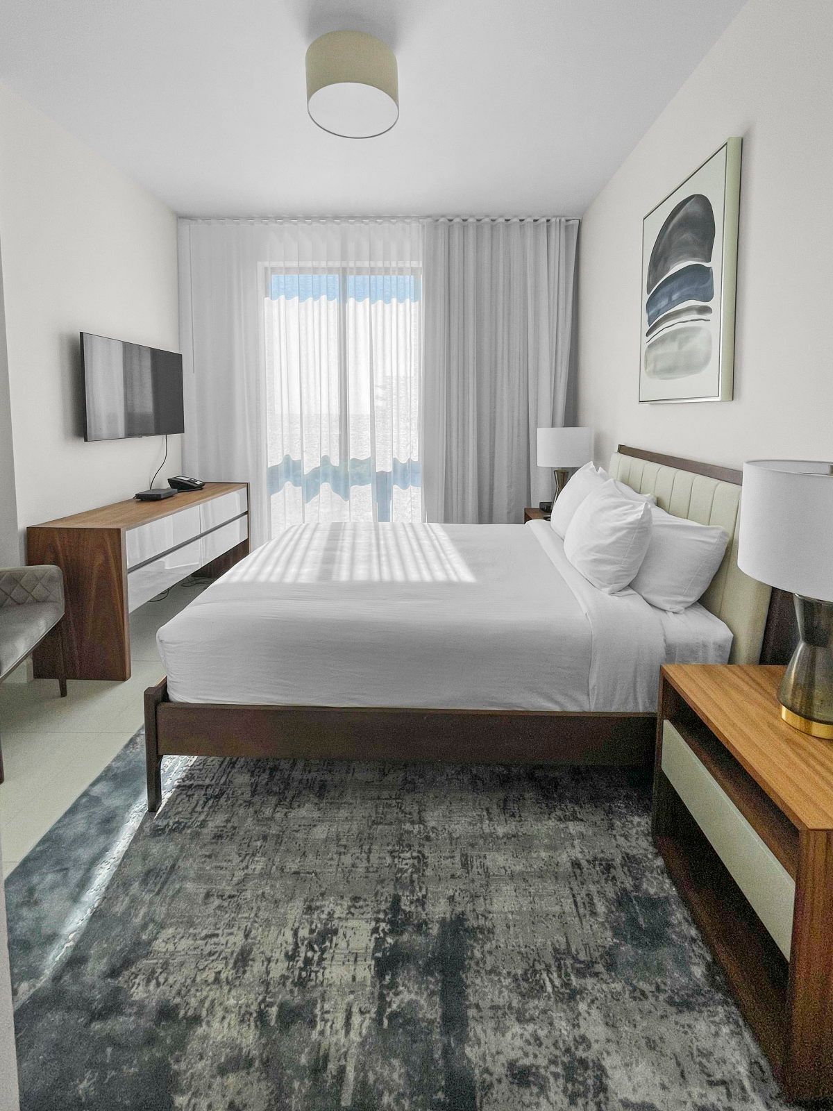 Ein modernes Hotelzimmer mit Bett, Nachttischen, Fernseher, Schreibtisch und einem großen Fenster mit Vorhängen.