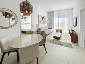 Moderni hotellihuone, jossa on ruokailutila, sohva, televisio ja taideteoksia seinillä.