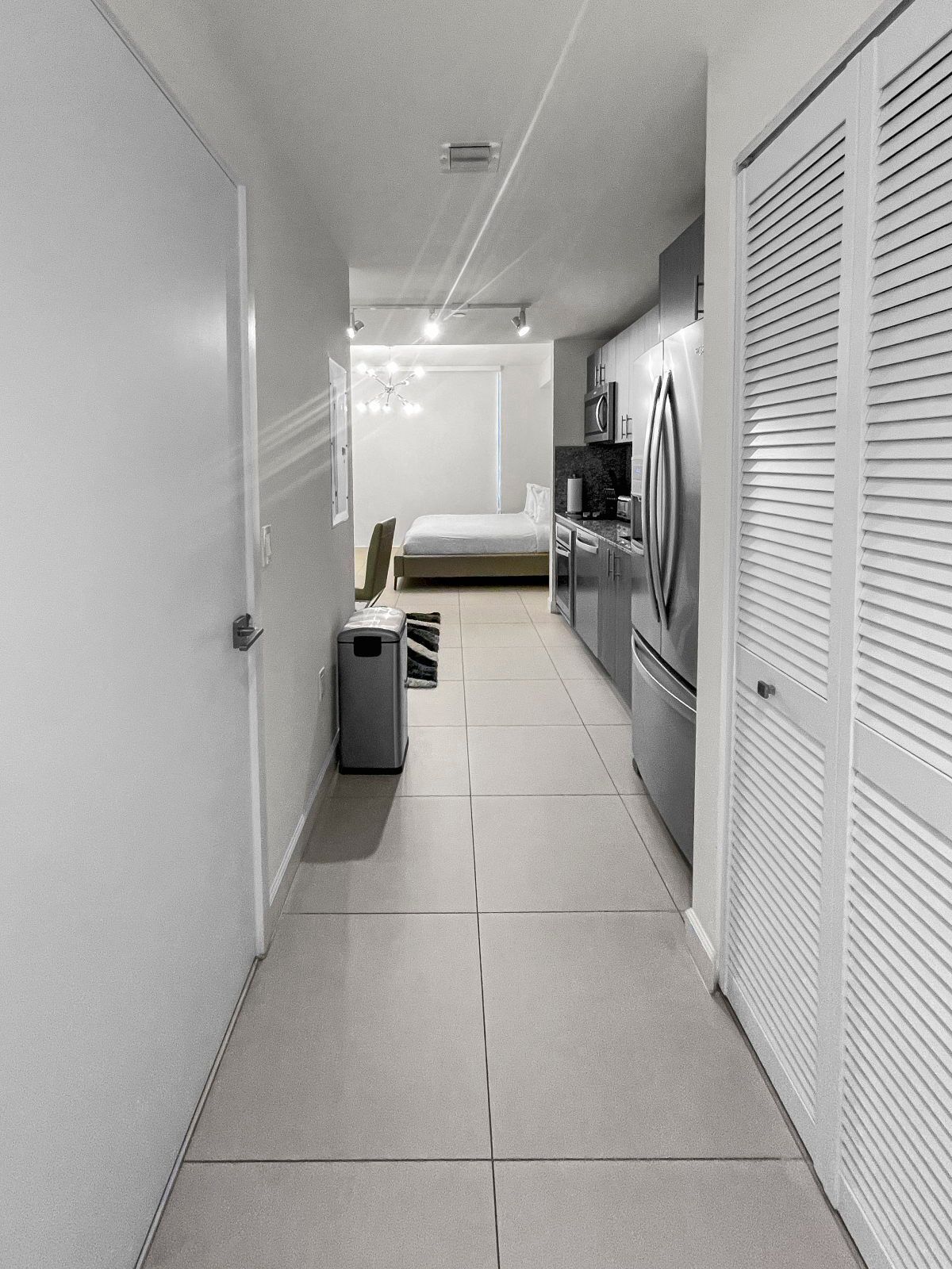A narrow hallway with a bed, kitchenette, and a trash bin.