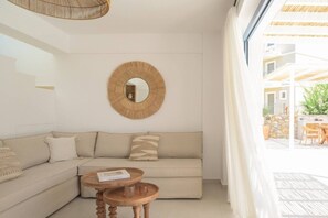 A modern living room with a sofa, two wooden side tables, a round mirror, and a view of an outdoor patio. - New Villa W/Heated Pool, 5 min Walk to Beach & Amenities (Georgioupoli)