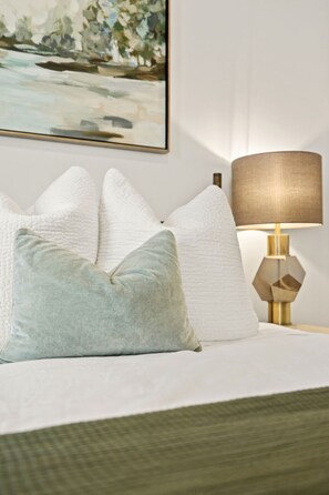 Uma cama com almofadas brancas e azul claras, uma luminária dourada e um quadro na parede.