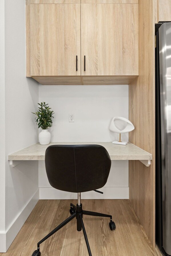 Eine moderne Küche mit Holzausstattung, einer weißen Arbeitsplatte, einem schwarzen Bürostuhl, einer weißen Vase mit Pflanze und einer weißen Schreibtischlampe.