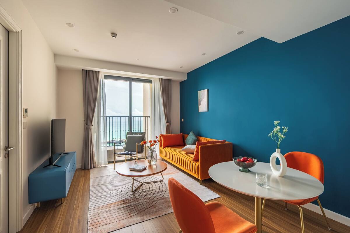 A modern living room with a sofa, coffee table, and chairs, a TV, and a balcony with curtains.