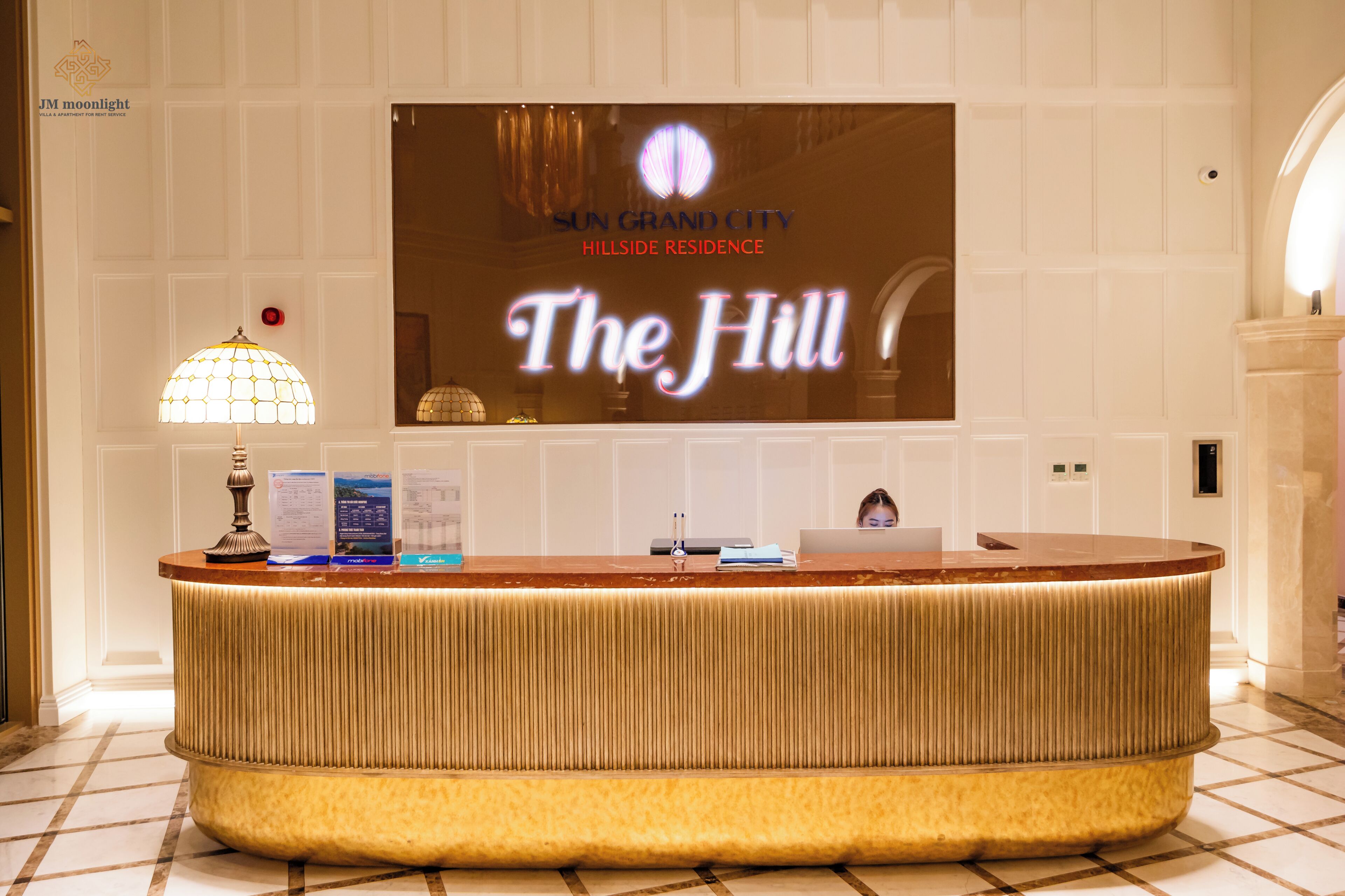 Een receptiebalie met een flatscreen-tv waarop het bord 'The Hill' staat, een lamp en informatiefolders.