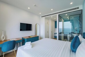 Et moderne hotelværelse med seng, skrivebord, stol, fjernsyn, spejl og badeværelse med glasvæg omkring bruseren.