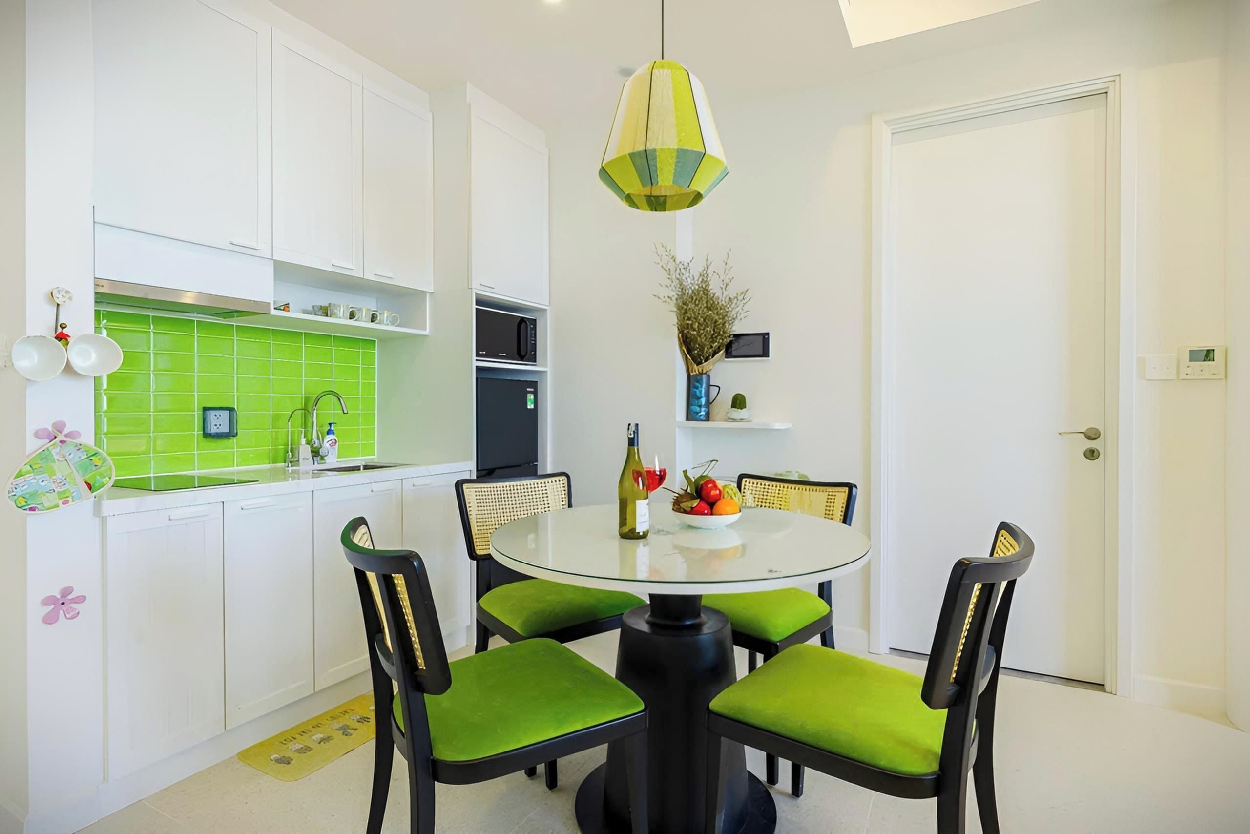A modern kitchen with white cabinets, a green backsplash, a round glass table with green chairs, and a hanging light fixture.