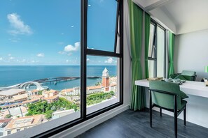 Ein Zimmer mit großem Fenster und Blick auf eine Küstenstadt, darunter eine Brücke und ein Turm.