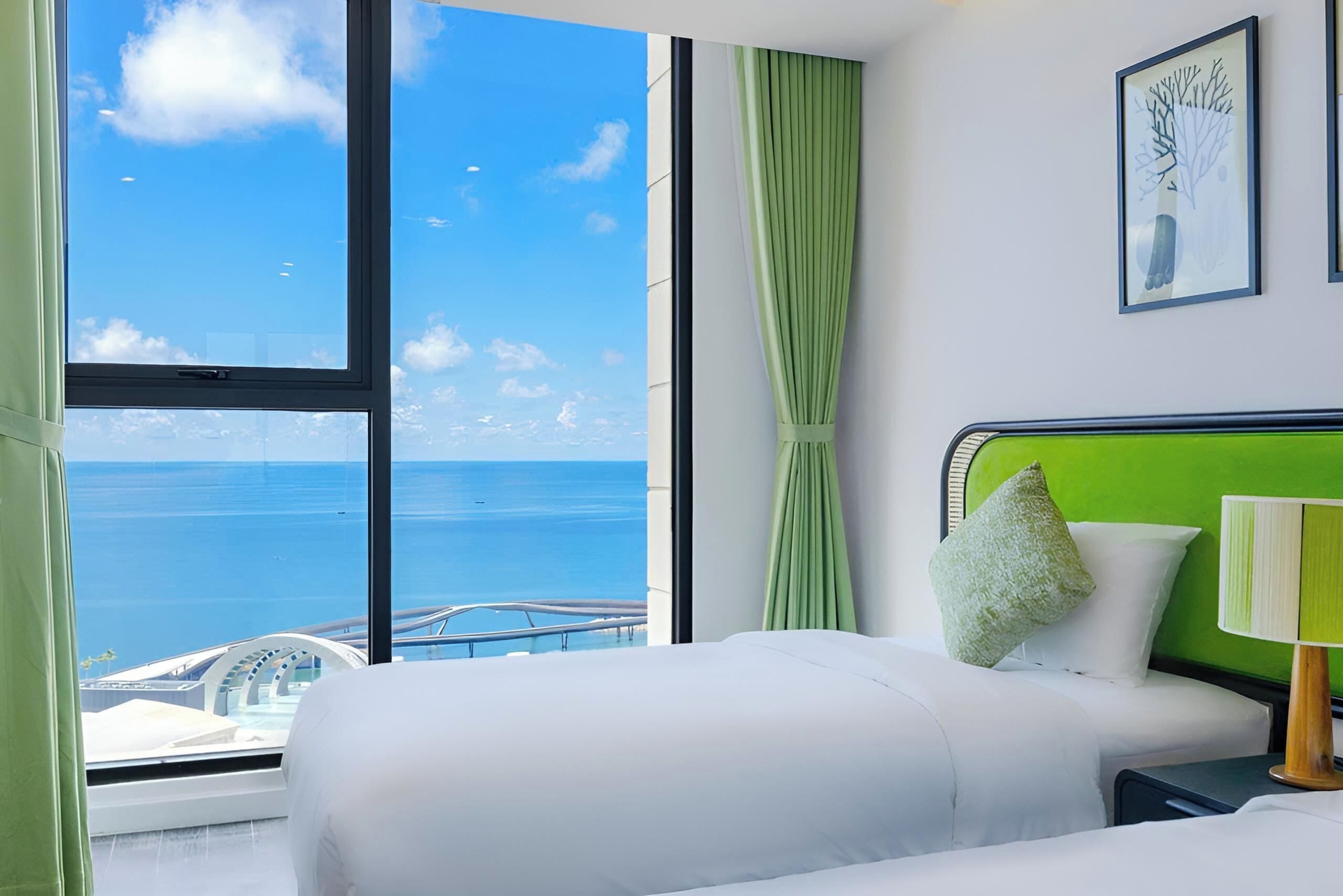 A hotel room with a large window offering a view of the sea and a bridge, a bed with white bedding, a green headboard, and a nightstand with a lamp.