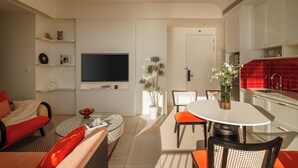 Una sala de estar moderna con televisor de pantalla plana, un comedor con mesa redonda y sillas, y una cocina con gabinetes blancos y azulejos rojos.