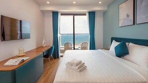 Habitación de hotel con cama, escritorio, silla, televisor y balcón con cortinas.