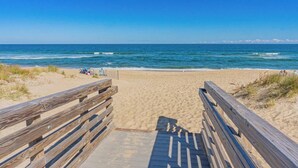 Ein hölzerner Steg führt zu einem Sandstrand, im Hintergrund das Meer.