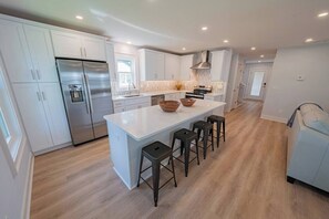 A modern kitchen with white cabinets, a stainless steel refrigerator, a large island with stools, and a built-in oven. - Return on Relaxation | Walk to Beach + Trails (Nags Head)
