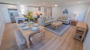 Eine moderne Küche mit weißen Schränken, ein Essbereich mit einem Holztisch für vier Personen und eine Wohnfläche mit zwei Sofas und einem Couchtisch.