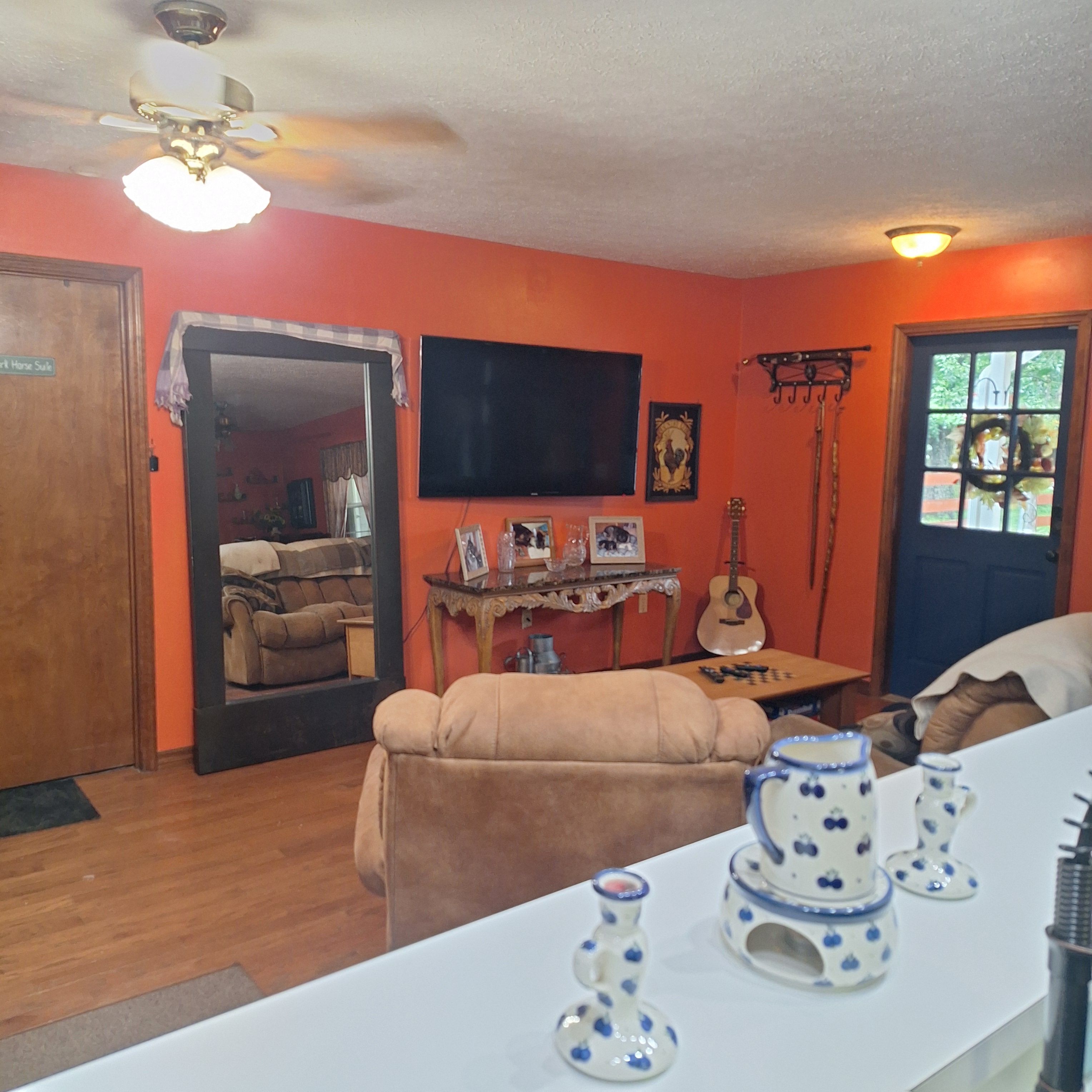 A room with a wooden floor, a beige sofa, a television mounted on the wall, a guitar, and a window with a wreath.