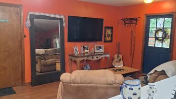 A room with a wooden floor, a beige sofa, a television mounted on the wall, a guitar, and a window with a wreath.