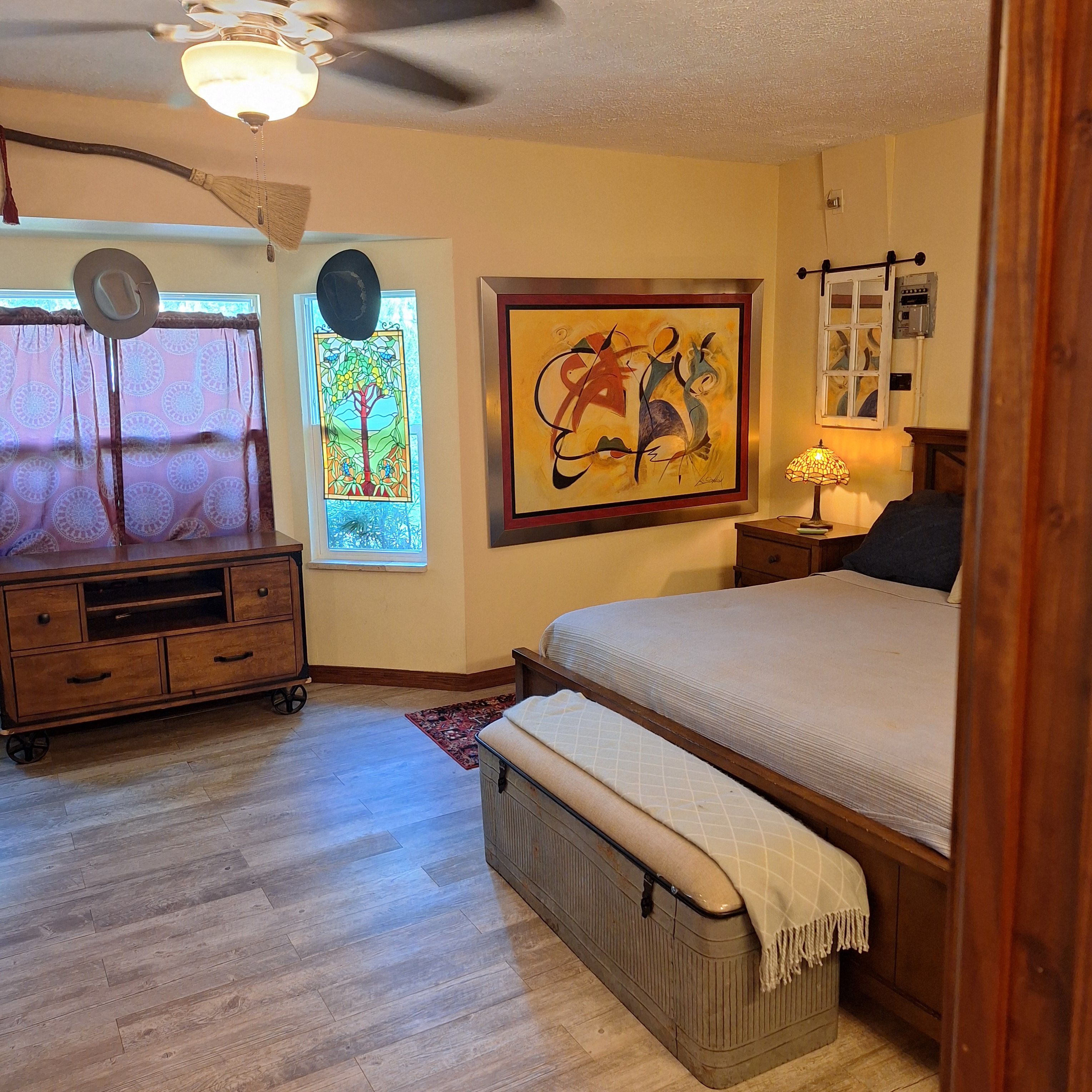A bedroom with a bed, a dresser, a window with curtains, a ceiling fan, and a wall-mounted lamp.