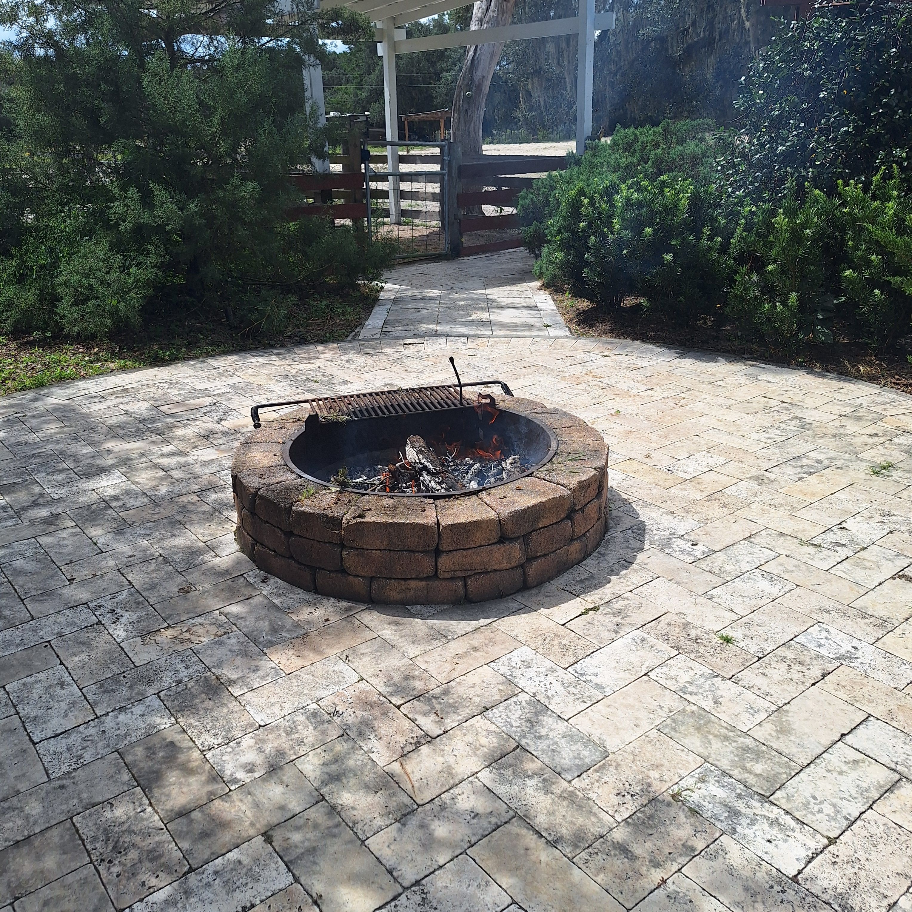 A circular fire pit with a metal grill, surrounded by a brick border, on a paved patio.