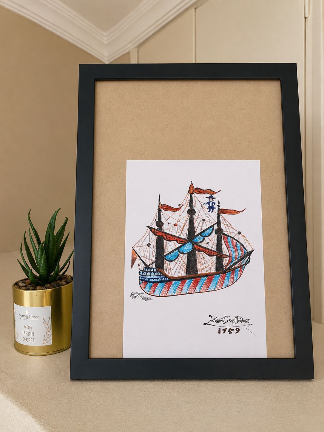 Una obra de arte enmarco que muestra un barco con el año 1759, una pequeña planta en maceta y un recipiente dorado con texto.