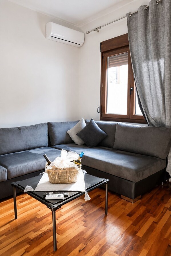 Un salon moderne avec un canapé gris, une petite table sur laquelle se trouve un panier, et une fenêtre avec des rideaux.