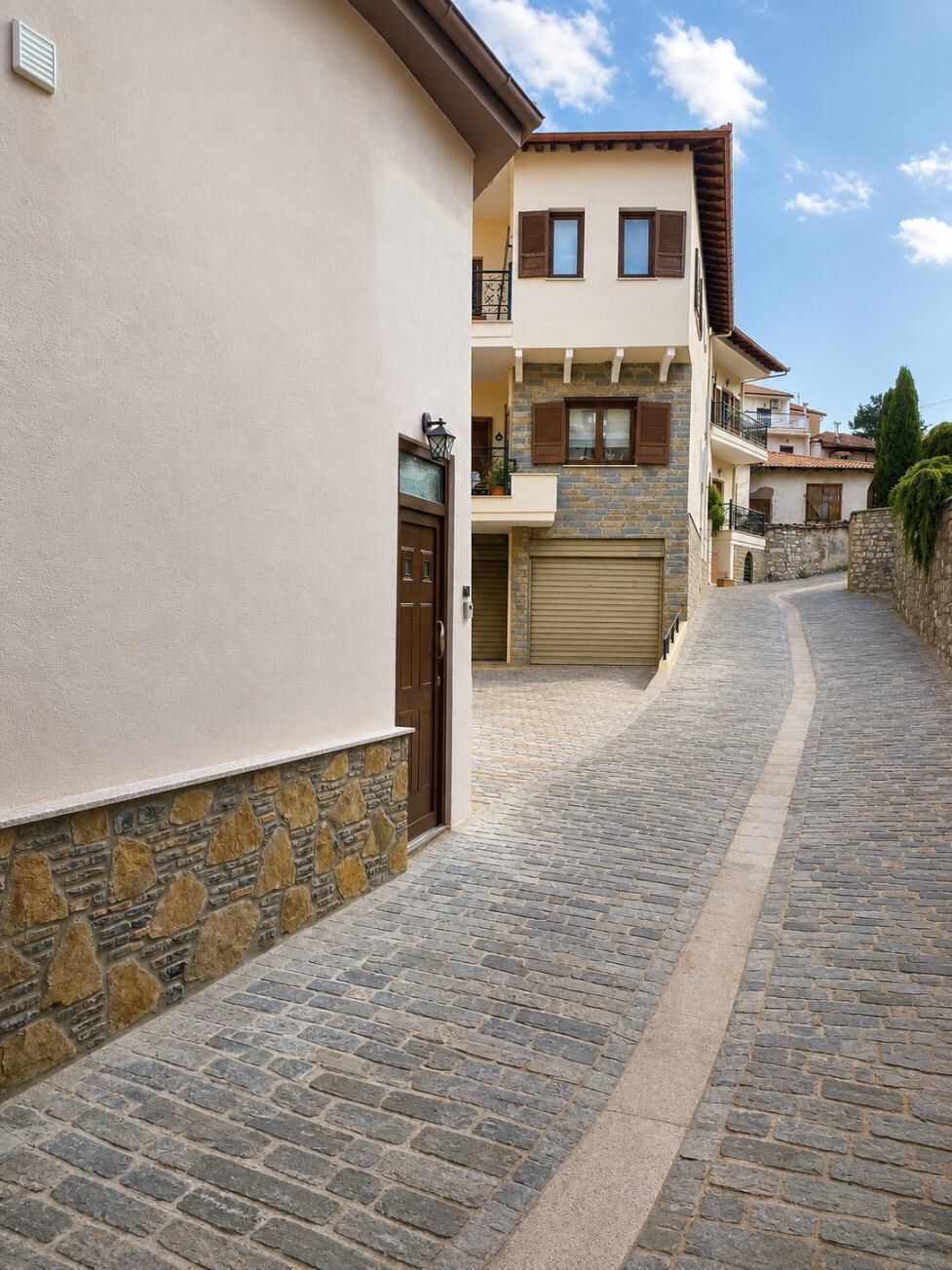 Una estrecha calle adoquinada flanqueada por edificios con fachadas de piedra y enlucido, una puerta de garaje cerrada y un cielo despejado.