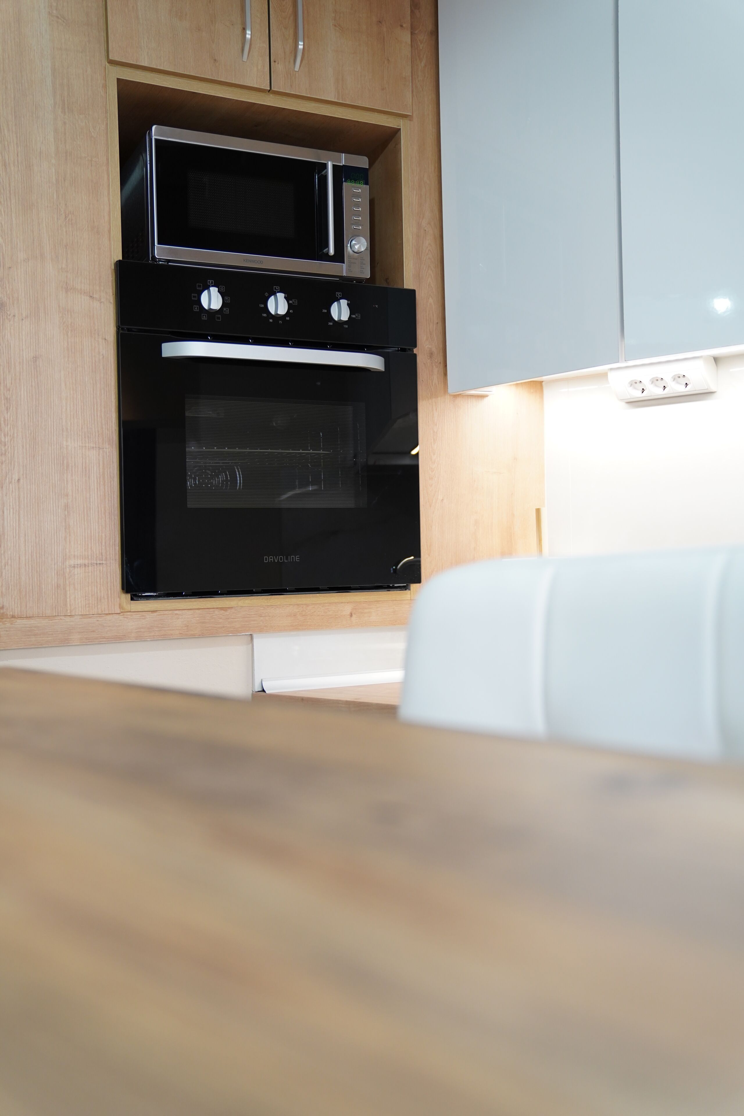 Une cuisine équipée d’un four et d’un micro-ondes encastrés, de meubles en bois et d’un plan de travail blanc.