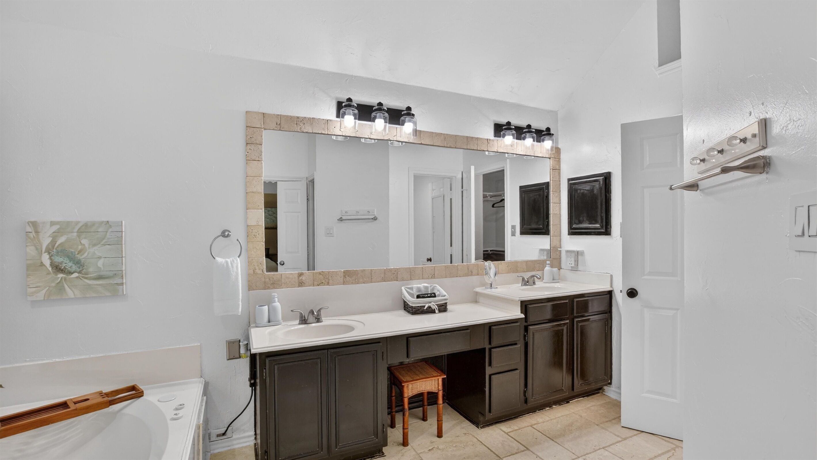A bathroom with a large mirror, two sinks, dark cabinets, a wooden stool, and a towel rack.