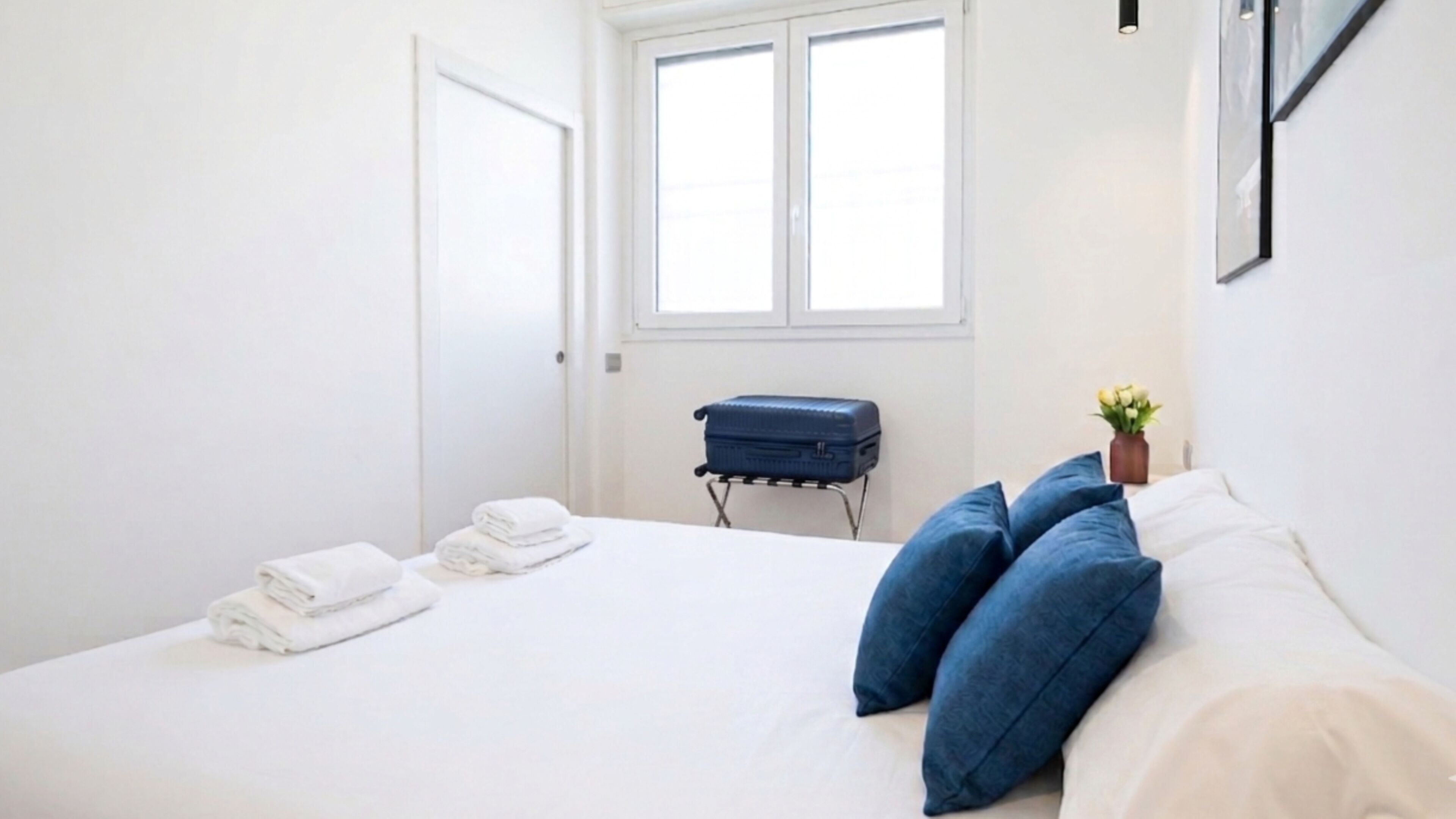 Una habitación de hotel con una cama, almohadas, una maleta y una ventana.