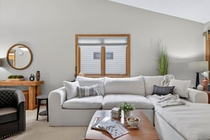 Una sala de estar moderna con un sofá modular, una mesa de centro de madera, un espejo redondo y una ventana con persianas.