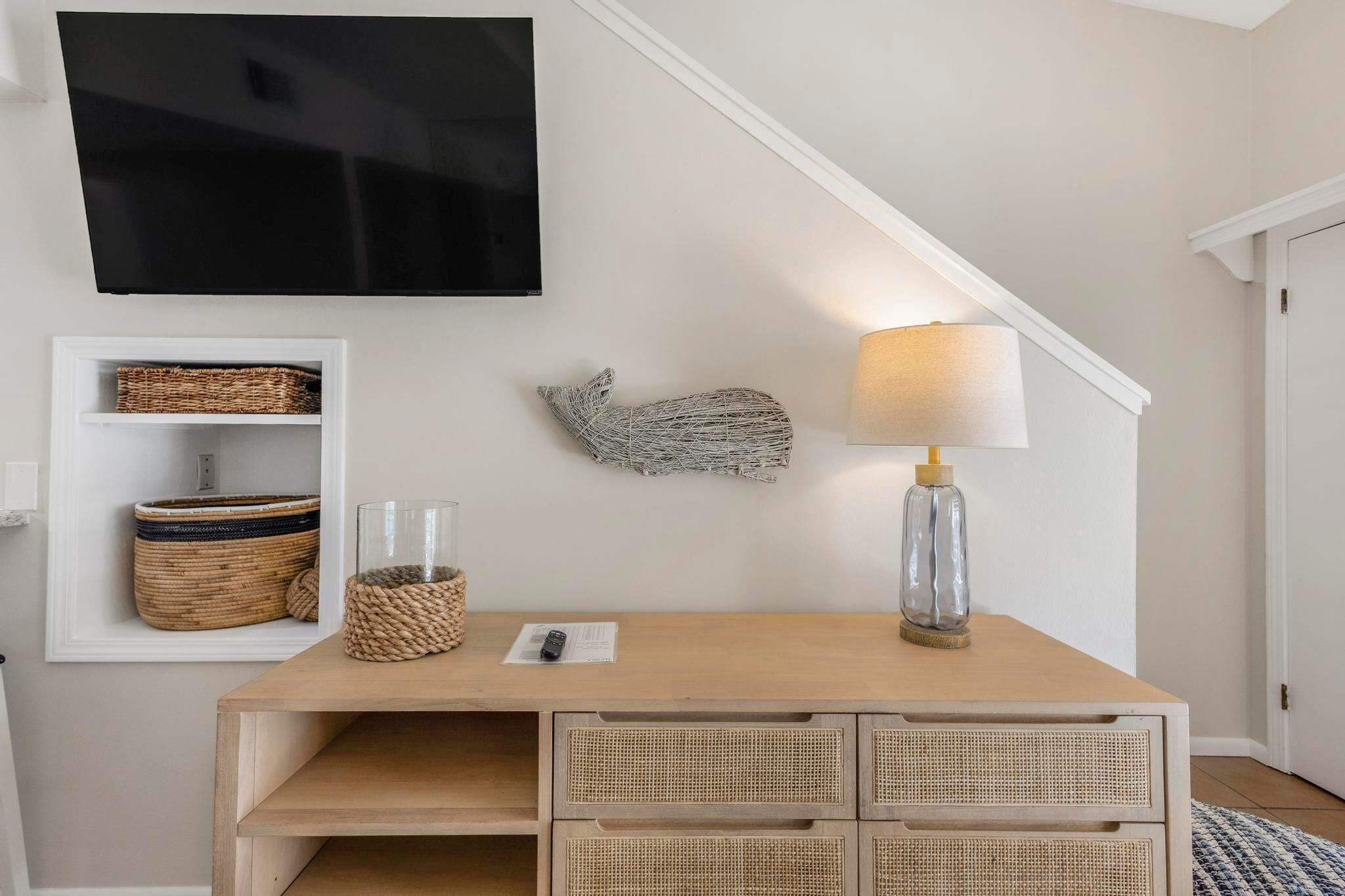 Ein Zimmer mit an der Wand montiertem Fernseher, einem hölzernen Schreibtisch mit Schubladen, Weidenkörben und einer Lampe.