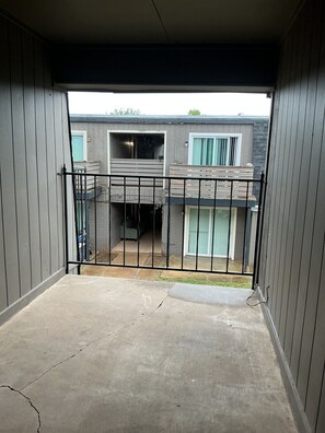 Blick auf einen eingezäunten Außenbereich mit Betonboden, einem Metallgeländer und einem Gebäude mit mehreren Fenstern und einem Balkon.
