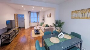 A modern living room with a sofa, a dining table with chairs, a TV, and a window with curtains. - Luxury 3BR Home 20 Mins to London Garden Wi-fi (Gravesend)