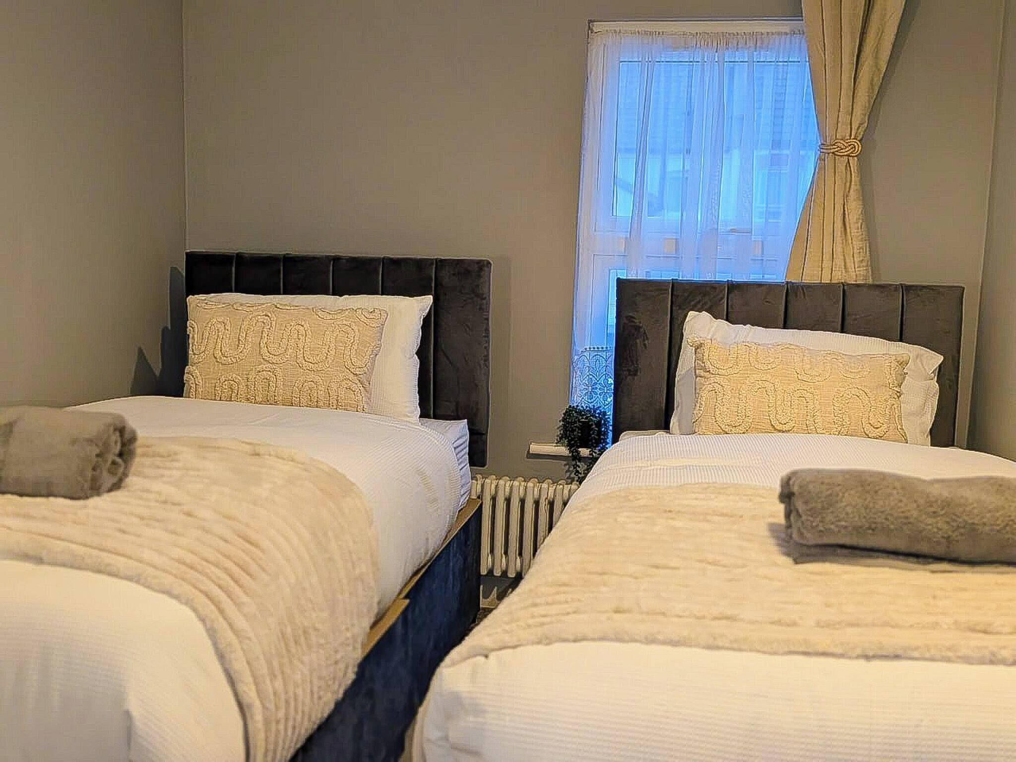 A hotel room with two beds, each with a headboard, a radiator, and a window with curtains.