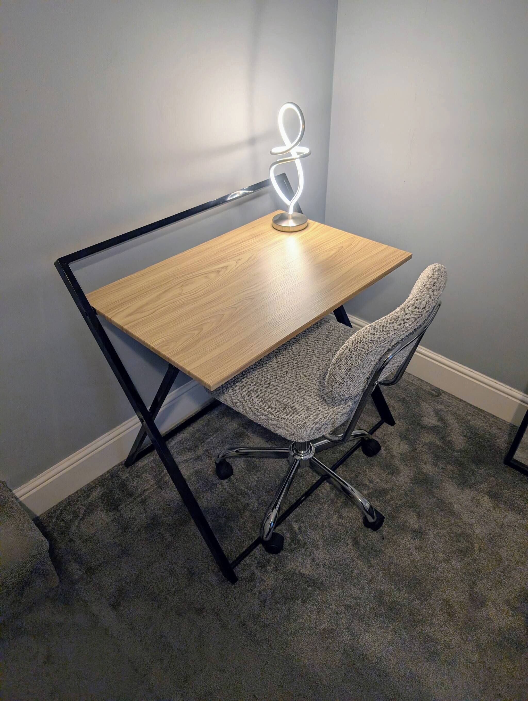 A corner desk with a wooden top and metal frame, a grey office chair with wheels, and a desk lamp with a circular light.