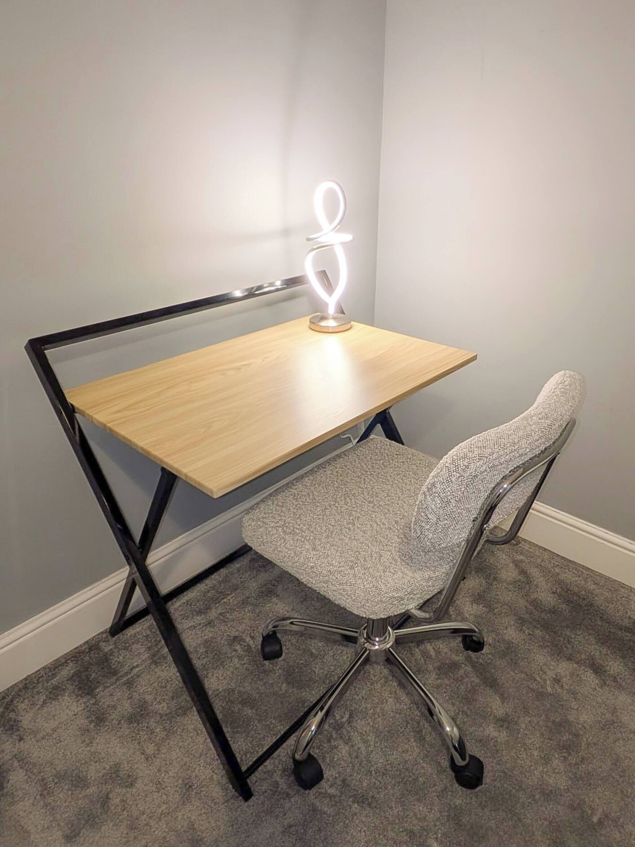 Ein Schreibtisch im Eckbereich mit einer beleuchteten Schreibtischlampe, ein grauer Bürostuhl und ein Teppichboden.