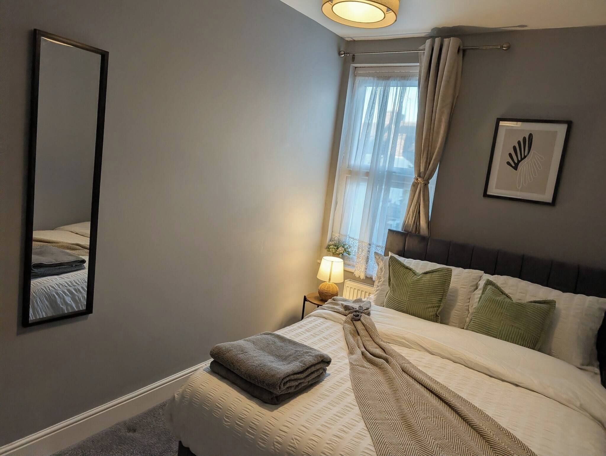 A bedroom with a bed, a nightstand, a lamp, a window with curtains, a framed picture, and a mirror.